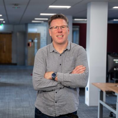 Chris stands in one of the team workspaces in the ONS open plan offices with his arms crossed smiling. He wears a relaxed grey flannel shirt and jeans. In the background there is a glass walled office and sit/stand desks.