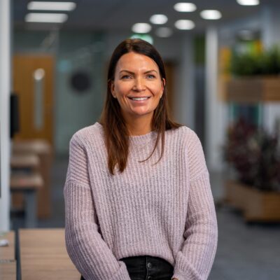 Jenny stands in one of the team workspaces in the ONS open plan offices with her hands held gently to her front, she's smiling and wears a grey knitted jumper and black jeans.
