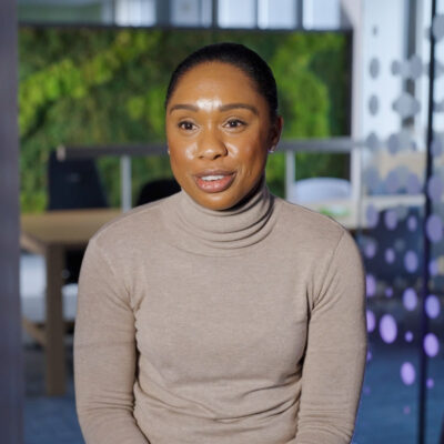 Keesia sits in one of the team workspaces in the ONS open plan offices with her hands held gently to her front, she's smiling and wears a grey turtle neck jumper.