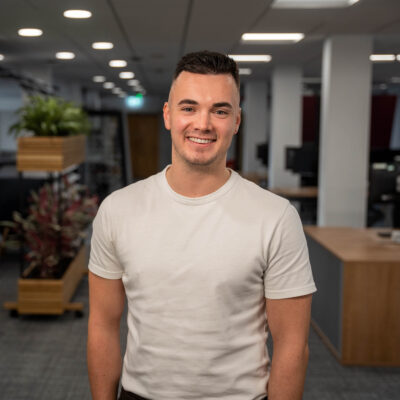 Jack stands in the centre of the open plan office space smiling. He wears a white T-shirt.