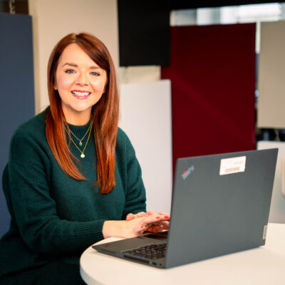 Rachel sits in front at a desk with her laptop. She's looking directly at the viewer and smiling and wears a forest green wool jumper.