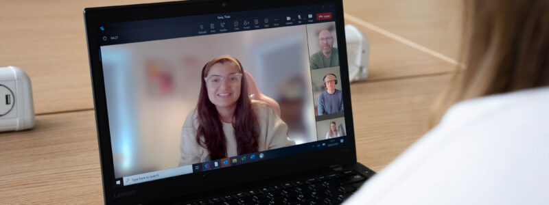 A view over the shoulder of a person who is engaged in a Teams video call on a laptop at an office desk. A young woman is presenting in full screen and is smiling with three other people on the call.